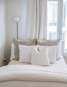 Cama ordenada con frazadas dobladas, almohadas, sábana blanca, ventana para luz natural, conectores en la pared