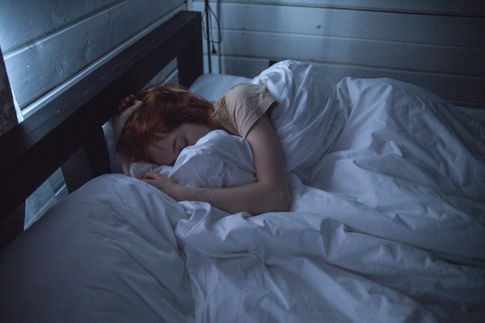 Mujer pelirroja durmiendo cómodamente en una cama con frazadas y almohadas blancas