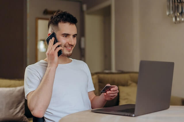 Adulto joven con tarjeta en mano para financiar muebles usando su laptop mientras habla por teléfono